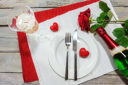 Table setting for a Valentines dinner, red rose and two hearts, champagne glass and bottle around a white plate on napkins and on a rustic wooden table, love concept, high angle view from aboveの写真素材