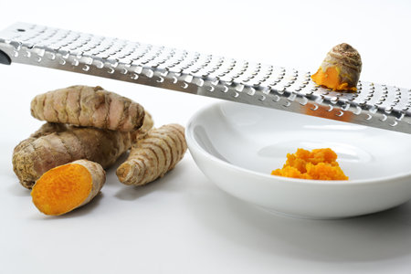 Turmeric roots are grated on a metal grater into a small white bowl for a healthy and spicy meal, light background, selected focus, narrow depth of fieldの写真素材