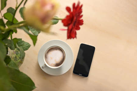 Coffee cup and mobile phone under a flower bouquet on a light wooden table, business concept for constant accessibility, copy space, top view from above, selected focus, narrow depth of fieldの写真素材