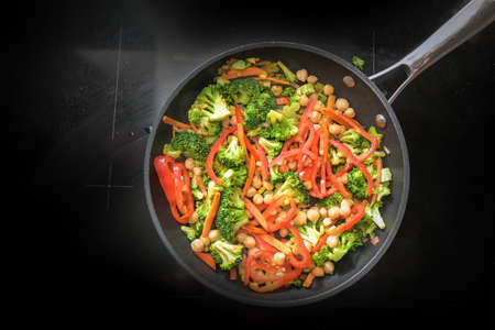Vegetable pan with broccoli, red bell peppers and chickpeas on a black stove, healthy cooking for a vegan low carb diet to lose weight, copy space, high angle view from aboveの写真素材