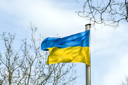 Flag of Ukraine with stripes in blue and yellow waving between trees against a cloudy sky, copy...
