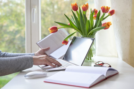 Hands of a woman typing on a laptop computer, books, mouse and tulip bouquet on a white desk with by...