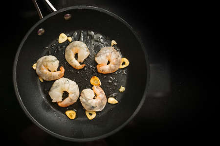 Fresh shrimp are sauteed with garlic in olive oil in a cooking pan on the black stove, preparation for a delicious appetizer, copy space, high angle view from above, selected focusの写真素材
