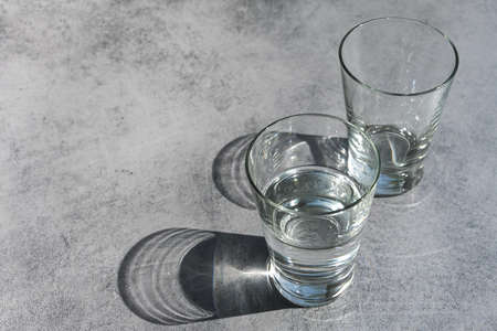 Drinking glasses, one half filled with water, the other empty, with shadows and light reflections on a rustic gray background, concept for climate change and water shortage, copy space, selected focusの写真素材