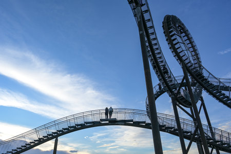 Couple standing on the stairs of the walkable roller coaster sculpture Tiger and Turtle against a blue sky with white clouds, tourist destination, art installation and landmark in Duisburg, Germanyのeditorial素材