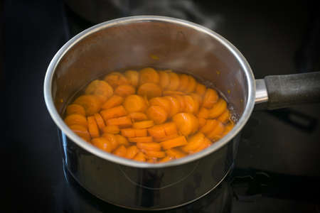 Slices of carrot are cooked in water in a steel pot on a black stove for a healthy digestible vegetable meal or soup against diarrhea, selected focus, narrow depth of fieldの写真素材