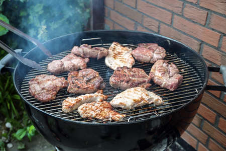 Steaks of various meats on a mobile round grill are roasted over charcoal at a barbecue party in the backyard, outdoor cooking concept, copy space, selected focus, narrow depth of fieldの写真素材