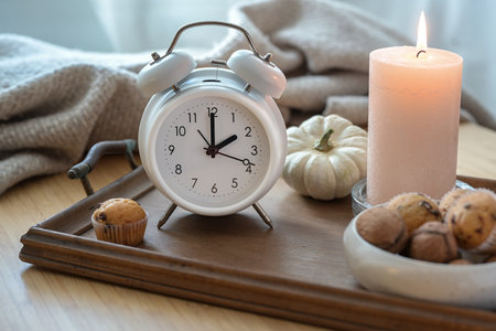 White vintage alarm clock, candle, autumn decoration and sweets on a nightstand with a natural woolen blanket, fall back concept after daylight saving time, selected focusの写真素材