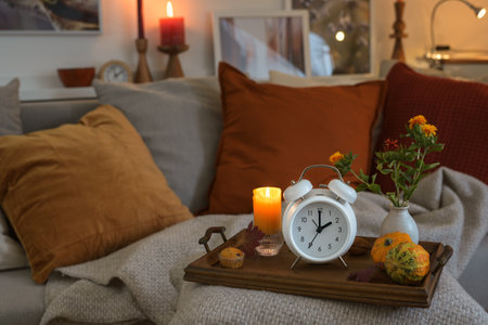 White vintage alarm clock showing fall back hour in autumn after daylight saving time, wooden tray with candle and warm colored fall decoration on a cozy couch, copy space, selected focusの写真素材