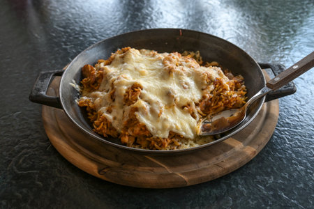 Fried rice with spices and topped with cheese in an iron pan on a wooden cutting board and a black table, fusion food, selected focus, narrow depth of fieldの写真素材