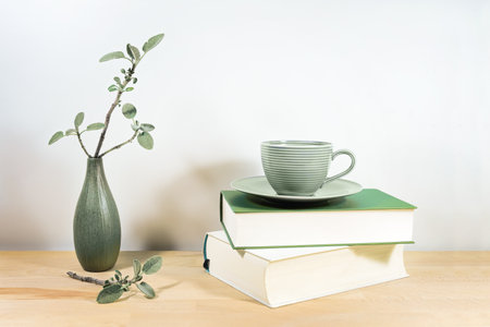 Wooden desk or table with books, a green tea cup and sage twigs in a small ceramic vase against a gray white wall, copy space, selected focus, narrow depth of fieldの写真素材