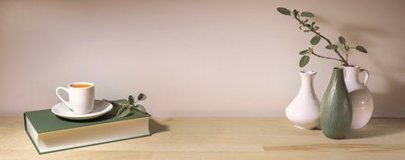 Coffee cup on a gray green book and a group of vases with a sage leaf branch on a wooden table against a pale wall, panoramic format, home decor, copy space, selected focusの写真素材