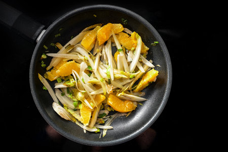 Slices of chicory and orange with spring onions in a black frying pan on the cooker to a vegetarian and vegan meal for a healthy low carb diet, high angle view from above, copy space, selected focusの写真素材