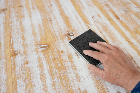 Hand with a sanding sponge removing white paint from the wooden surface of a large table, homemade...