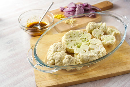 Raw cauliflower slices in a glass casserole, chopped red onion and a spice mix in olive oil, cooking preparation for baked vegetable steaks, copy space, selected focus, narrow depth of fieldの写真素材