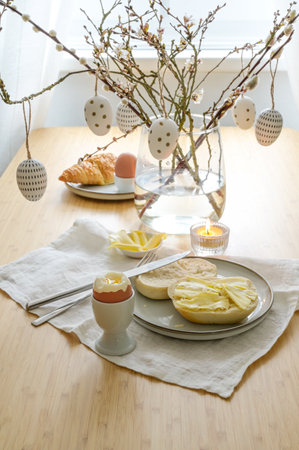 Easter breakfast with egg, bread roll and butter, decorated with spring branches and hanging patterned easter eggs in a glass vase on a dining table, copy space, selected focus, narrow depth of fieldの写真素材