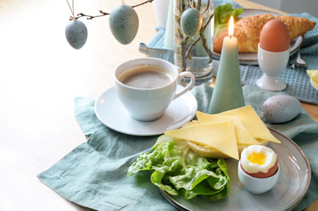 Breakfast table setting with bread roll, cheese, boiled egg and coffee on a pastel turquoise napkin, hanging Easter eggs and candle as decoration, copy space, selected focus, narrow depth of fieldの写真素材