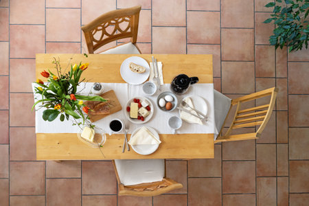 Breakfast on a wooden table with bread, cheese, eggs and coffee and a bouquet of tulips as decoration, three vintage chairs on a tiled floor, high angle view from above, selected focusの写真素材