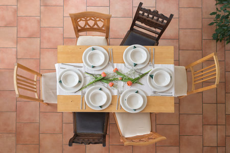 Wooden table setting with six empty white plates and bowls, cutlery and some tulip flowers, different vintage chairs around on a tiled terracotta floor, high angle view from above, selected focusの写真素材