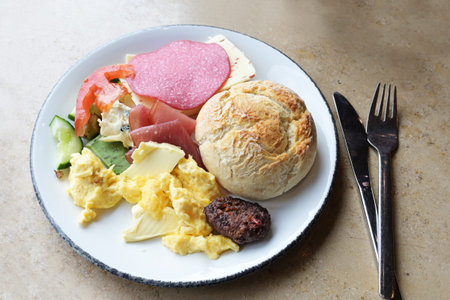 Breakfast plate with bread rolls, scrambled eggs, sausage, cheese and butter with cutlery on a stone...