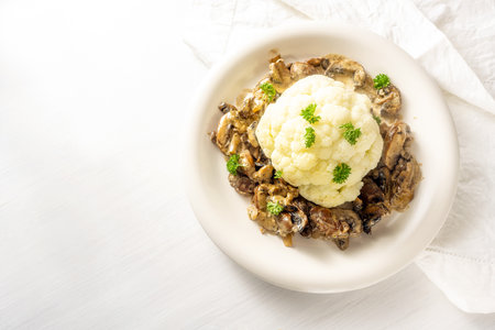 Cauliflower with mushroom ragout and parsley garnish on a plate and on a white table, healthy vegetable meal, high angle view from above, copy space, selected focus, narrow depth of fieldの写真素材