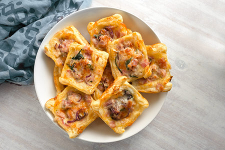Baked puff pastry squares with ham, onion, herbs and cheese in a bowl, finger food snack for a warm...