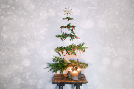 Christmas tree alternative, evergreen branches with decoration hanging on the wall above a rustic wooden stool with candles, sustainable holidays against a snowy background, copy space, selected focusの写真素材