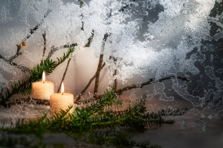 White burning candles with warm light and a fir tree branch against a frozen window glass, winter greeting card or Christmas decoration, copy space, selected focus, narrow depth of fieldの写真素材