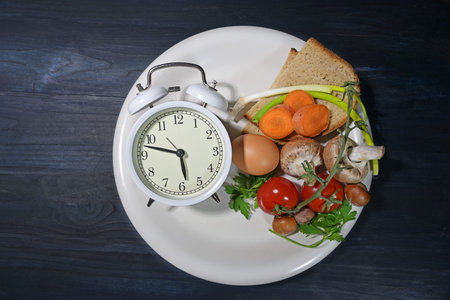 Intermittent fasting, white plate with an alarm clock and partly filled with food on dark blue wood,...