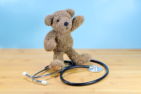 Little brown teddy bear standing on a stethoscope and waving, health care for children, pediatric medicine concept, blue background with copy space, selected focusの写真素材