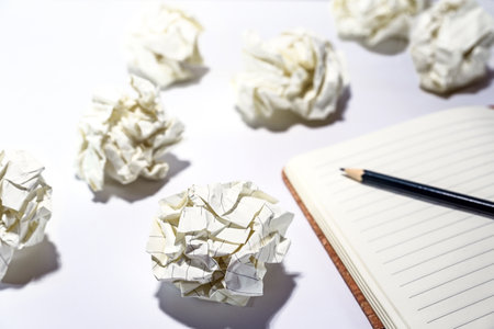Discarded crumpled paper balls around an empty notebook with a pencil, concept for creative writing, development process, business frustration and stress, white table, copy space, selected focusの写真素材
