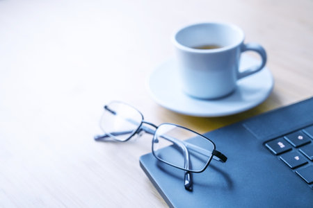 Part of a black laptop, glasses and a cup of coffee on a wooden desk in an office, concept for business, study, writing or journalism, copy space, selected focus, narrow depth of fieldの写真素材