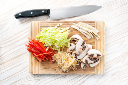 Chopped vegetables on a wooden cutting board with a kitchen knife, mushrooms, peppers, soybean sprouts and cabbage for cooking a vegetarian Asian meal, top view from above, selected focusの写真素材