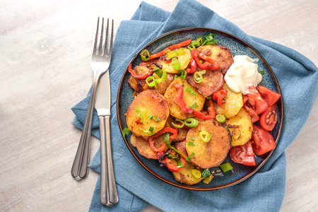 Fried potato slices, red bell pepper and green spring onions in a plate on a blue napkin and cutlery on a light painted wooden table, top view from above, copy space, selected focusの写真素材
