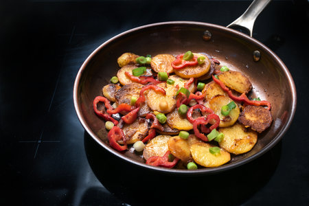 Crispy fried potatoes, red bell pepper and green spring onions in a frying pan on a black hob in the kitchen at home, cooking with vegetables, vegetarian meal, selected focus, narrow depth of fieldの写真素材