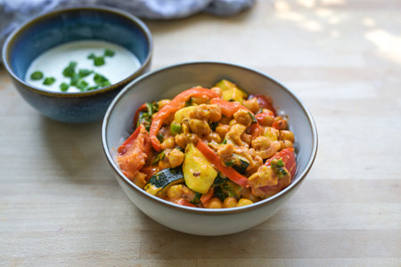 Vegetarian curry made from chickpeas and vegetables such as zucchini, peppers and leek in a bowl, served with fresh yogurt, healthy meal on a wooden table, copy space, selected focusの写真素材
