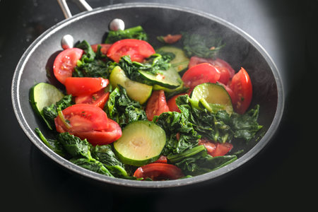 Steaming vegetables such as spinach, zucchini and tomatoes in a frying pan on a black stove top, cooking a healthy vegetarian meal at home, selected focus, narrow depth of fieldの写真素材