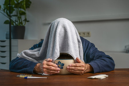 Elderly man with a towel over his head doing steam inhalation over a bowl with hot water at a table...