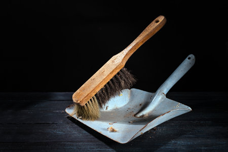 Old gray metal dustpan and brush with wooden handle against a very dark background, still life of vintage cleaning utensils, copy space, selected focusの写真素材