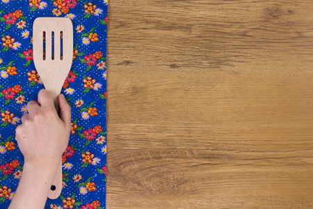Hand holding kitchen wood spatula.  Pattern flower cloth napkin on empty wooden background. Top view with space for your text.の写真素材