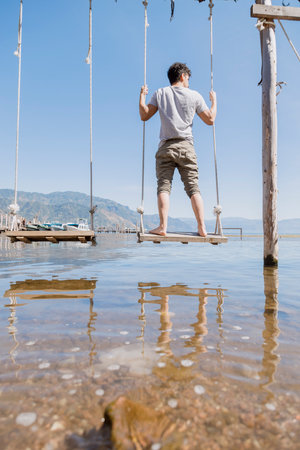 Young man standing on a swing in the middle of Lake Atitlan - Hispanic traveler enjoying Lake Atitlan in Guatemala - Lake landscape in the morningの写真素材