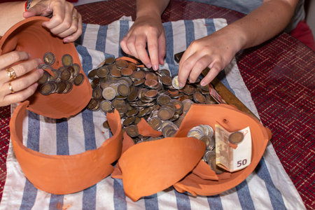 Four hands selecting coins from a piggy bank in the shape of a broken brown pig on a table - Piggy...