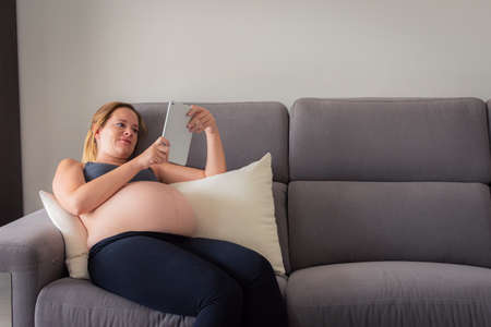 Pregnant Woman On Sofa Relaxing With Digital tablet reading some pregnancy tips. Woman with a 8 month of pregnancy reading on the sofa.の写真素材