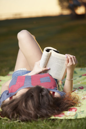 Smiling beautiful young woman lying on grass and reading book, against background of summer green...