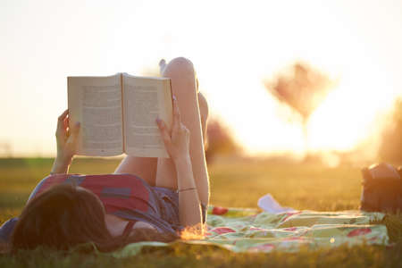 Smiling beautiful young woman lying on grass and reading book, against background of summer green parkの写真素材