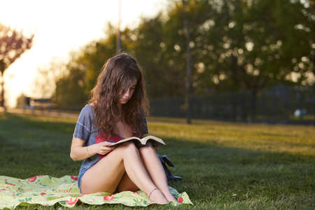 Smiling beautiful young woman lying on grass and reading book, against background of summer green parkの写真素材
