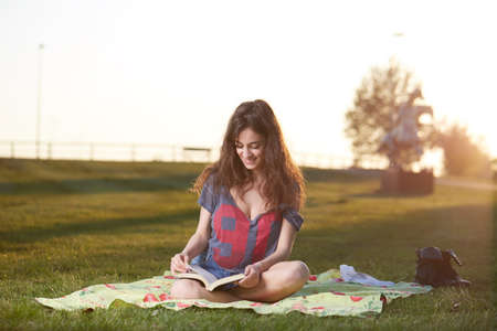 Smiling beautiful young woman lying on grass and reading book, against background of summer green parkの写真素材