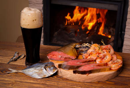Glass of dark beer and a tray of assorted fish on a background of a burning fireplaceの写真素材