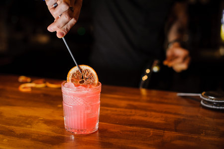 Bartender holding tongs in his hands adorns pink cocktail with an grapefruit slice. Barman at work,...