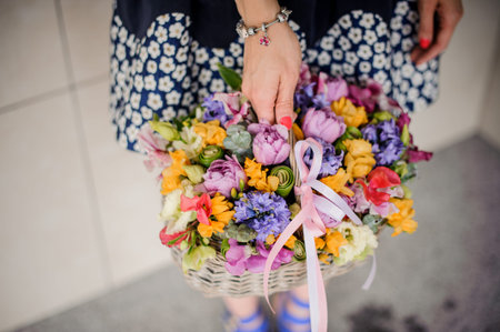 Woman holding pretty wicker basket of colorful flowers in hand on the light background no face close...